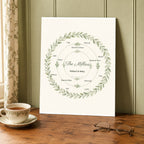 The unframed watercolor wreath print resting on a wooden table beside a teacup and reading glasses